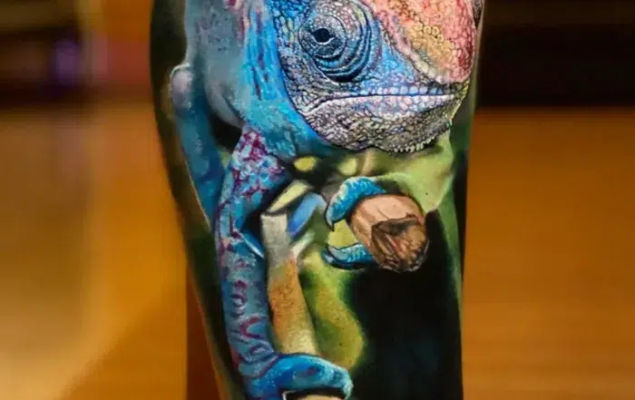 A highly detailed, realistic color tattoo on a person's leg depicting a vibrant chameleon. The chameleon is primarily bright blue with pink and orange highlights on its head, perched on a brown branch against a soft-focus green and dark background.