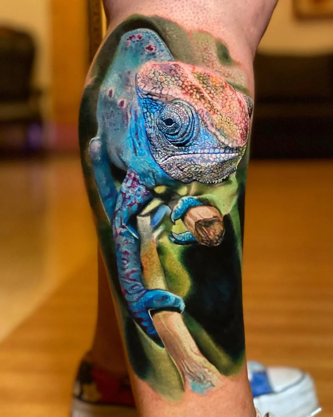 A highly detailed, realistic color tattoo on a person's leg depicting a vibrant chameleon. The chameleon is primarily bright blue with pink and orange highlights on its head, perched on a brown branch against a soft-focus green and dark background.