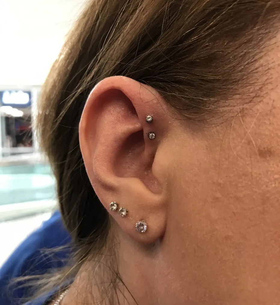 A close-up of a curated ear featuring a vertical double forward helix piercing with two small, matching clear gemstone studs. Three additional stud piercings of varying sizes descend along the earlobe, creating a cohesive, sparkling look.