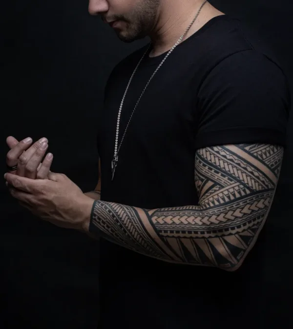 A bold, blackwork tattoo covering a man’s entire forearm and upper arm. The design consists of intricate, repeating geometric patterns and thick tribal bands with sharp lines and symmetrical shading.