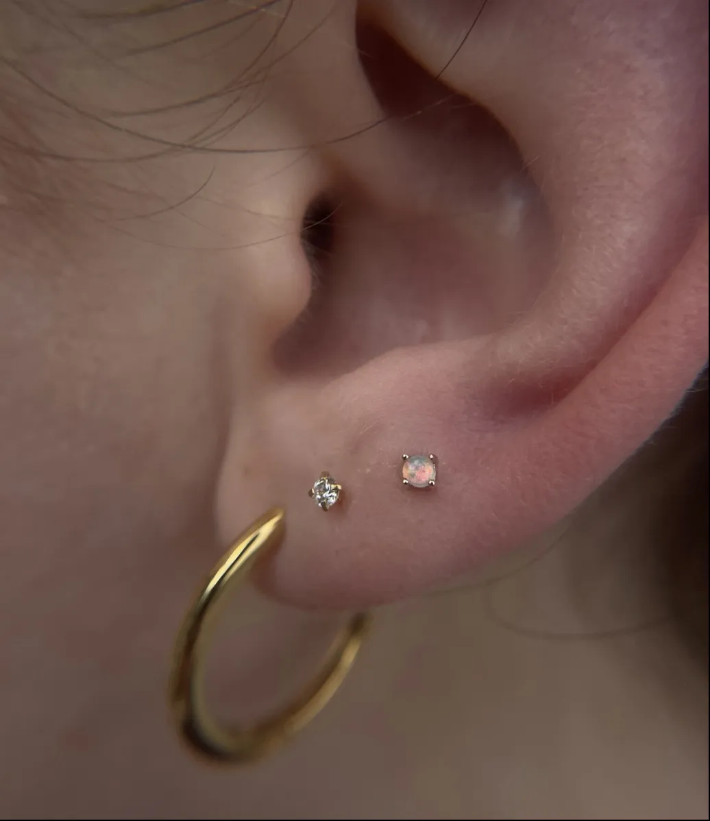A close-up of a person's ear featuring a classic earlobe piercing with a thick, polished gold hoop. Two additional small stud piercings are visible higher on the lobe: one featuring a clear diamond-like gem and the other a small, iridescent opal-style stone.