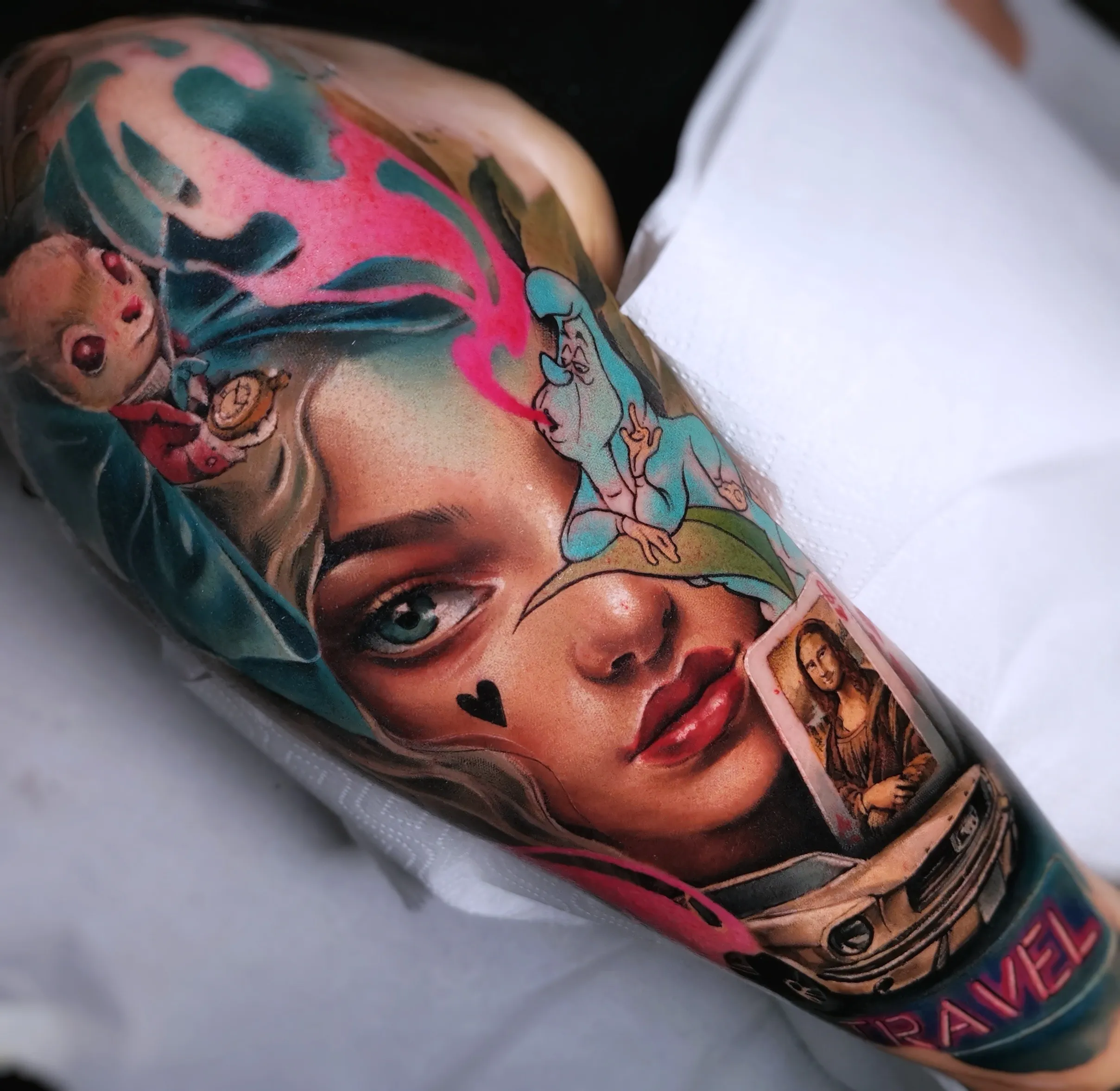 A complex, colorful surrealist tattoo on a shoulder. It features a realistic woman’s face with a small black heart on her cheek. Overlaid on the portrait are various "Alice in Wonderland" elements, including the White Rabbit with a pocket watch, a smoking blue caterpillar, a playing card with the Mona Lisa, and a classic car at the bottom with the word "TRAVEL."