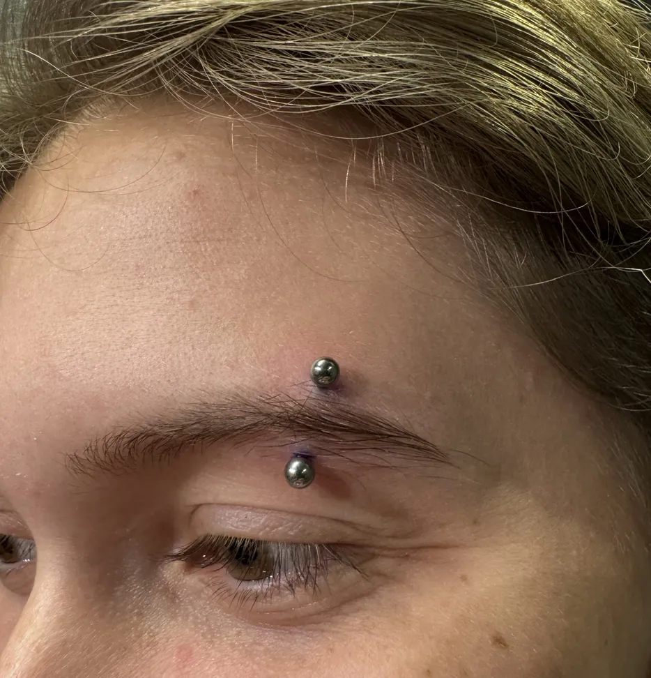 A close-up of a fresh eyebrow piercing on a person's right eye. The jewelry is a curved titanium barbell with two silver-colored metal spheres, positioned vertically through the outer tail of the eyebrow. Purple surgical marking ink is visible around the entry and exit points.