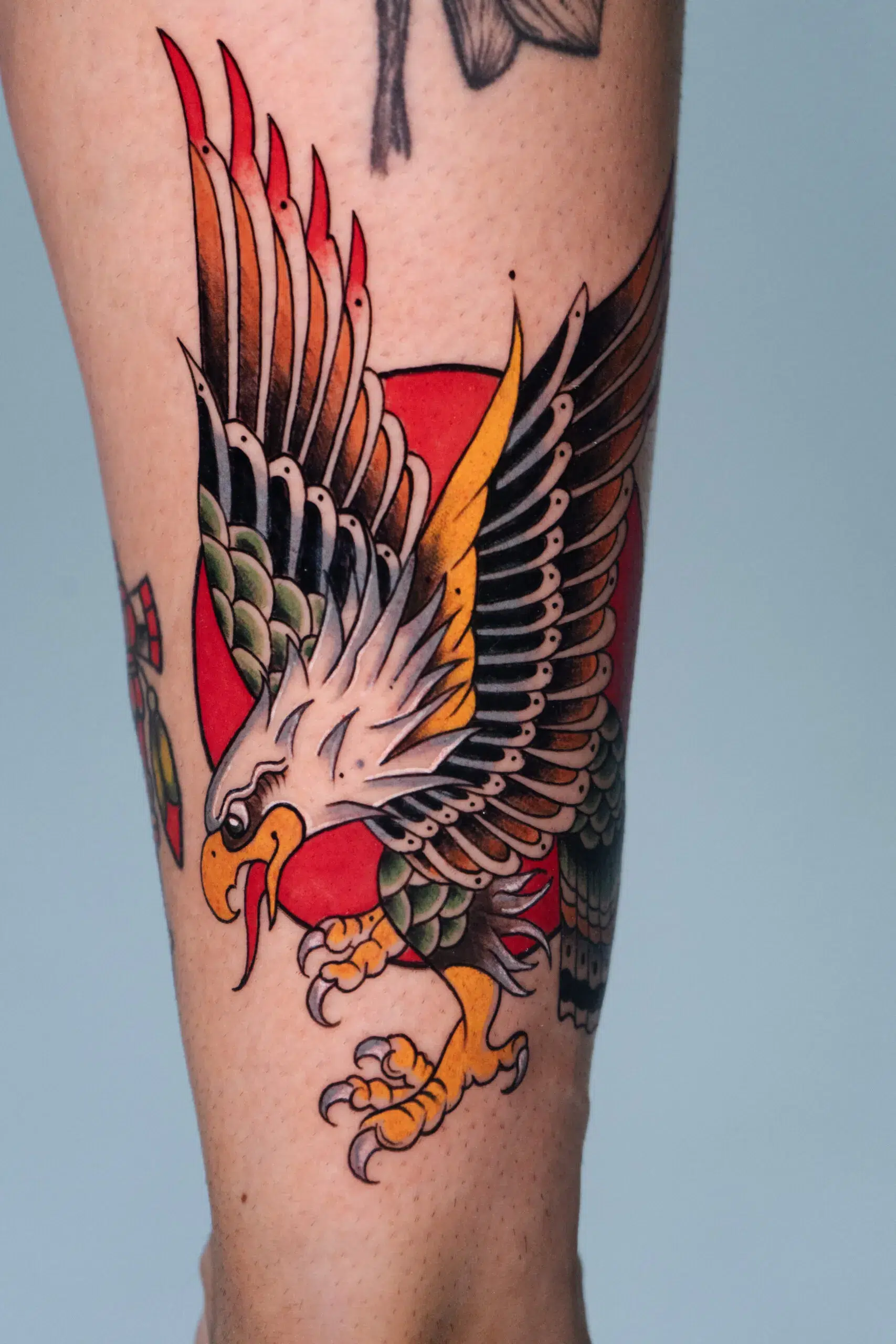 A classic American Traditional style tattoo on a forearm. It shows a powerful eagle in mid-flight with its talons extended, set against a large red sun. The eagle is rendered with bold black outlines and a palette of yellow, green, and brown.