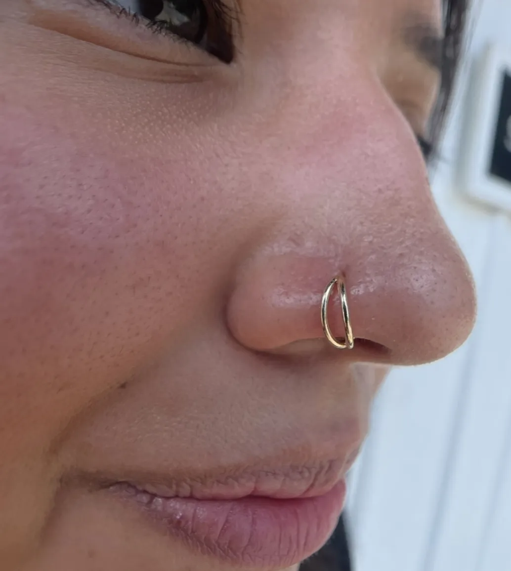 A side-profile view of a person’s nose featuring a delicate double-gold hoop nose ring. The jewelry consists of two thin, parallel gold wires passing through a single nostril piercing.
