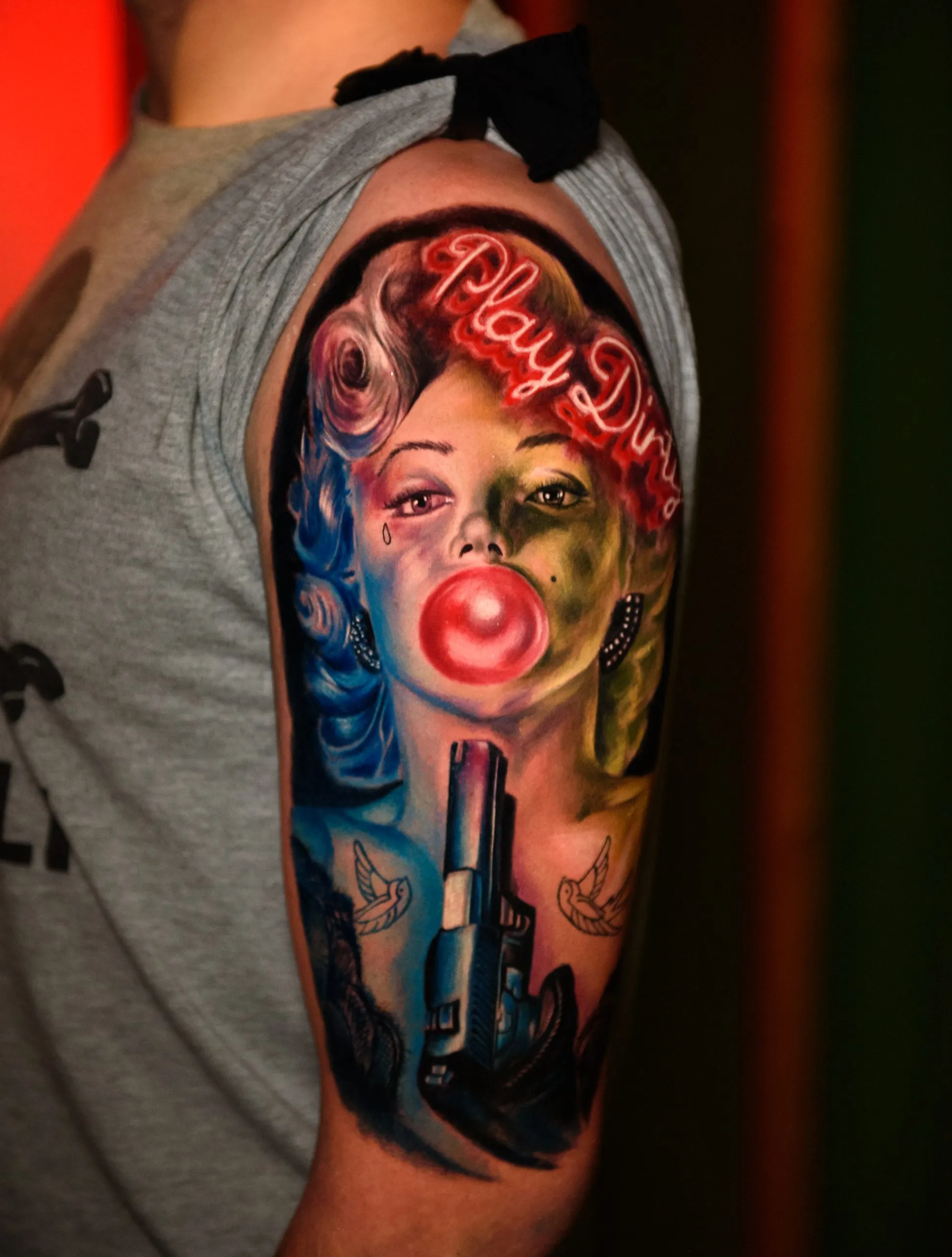 A stylized color portrait tattoo on a person’s upper arm. The image features a woman with blue and blonde hair blowing a pink bubble with bubblegum, set against a background with red neon-style text that reads "Play Dirty." A realistic handgun is positioned vertically in the foreground at the bottom of the portrait.