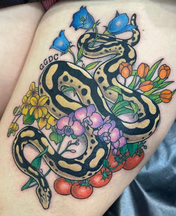 A vibrant color tattoo on a person's thigh featuring a yellow and black patterned ball python coiled among an assortment of flowers. The composition includes blue bellflowers, orange tulips, yellow pansies, purple orchids, and a cluster of red tomatoes at the bottom. The letters "GGDC" are tattooed in black near the top left of the design.