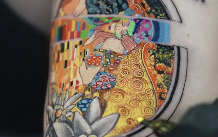 A detailed, circular micro-realism tattoo inspired by Gustav Klimt's "The Kiss." The art is encased in a geometric frame and features intricate patterns in gold, green, and red, with soft floral details at the bottom.