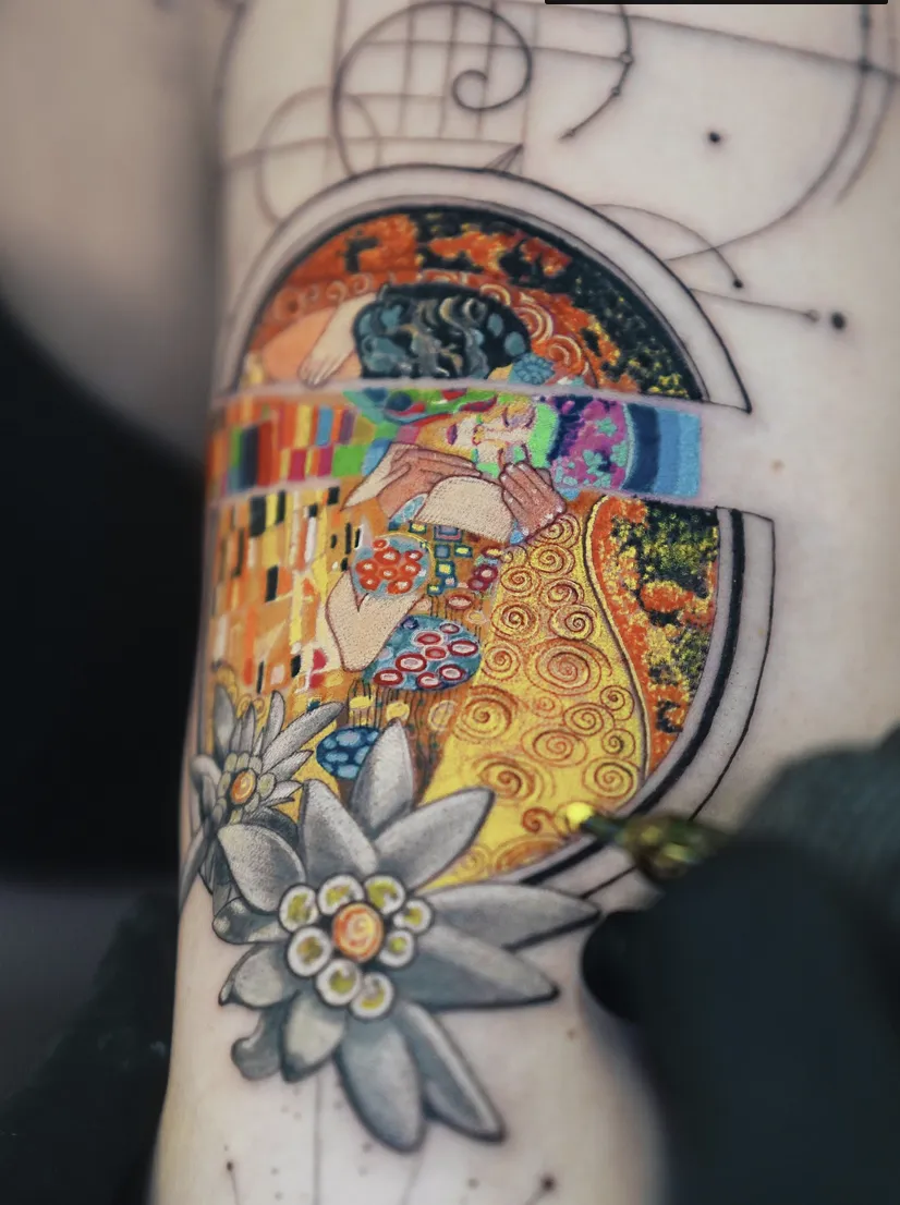 A detailed, circular micro-realism tattoo inspired by Gustav Klimt's "The Kiss." The art is encased in a geometric frame and features intricate patterns in gold, green, and red, with soft floral details at the bottom.