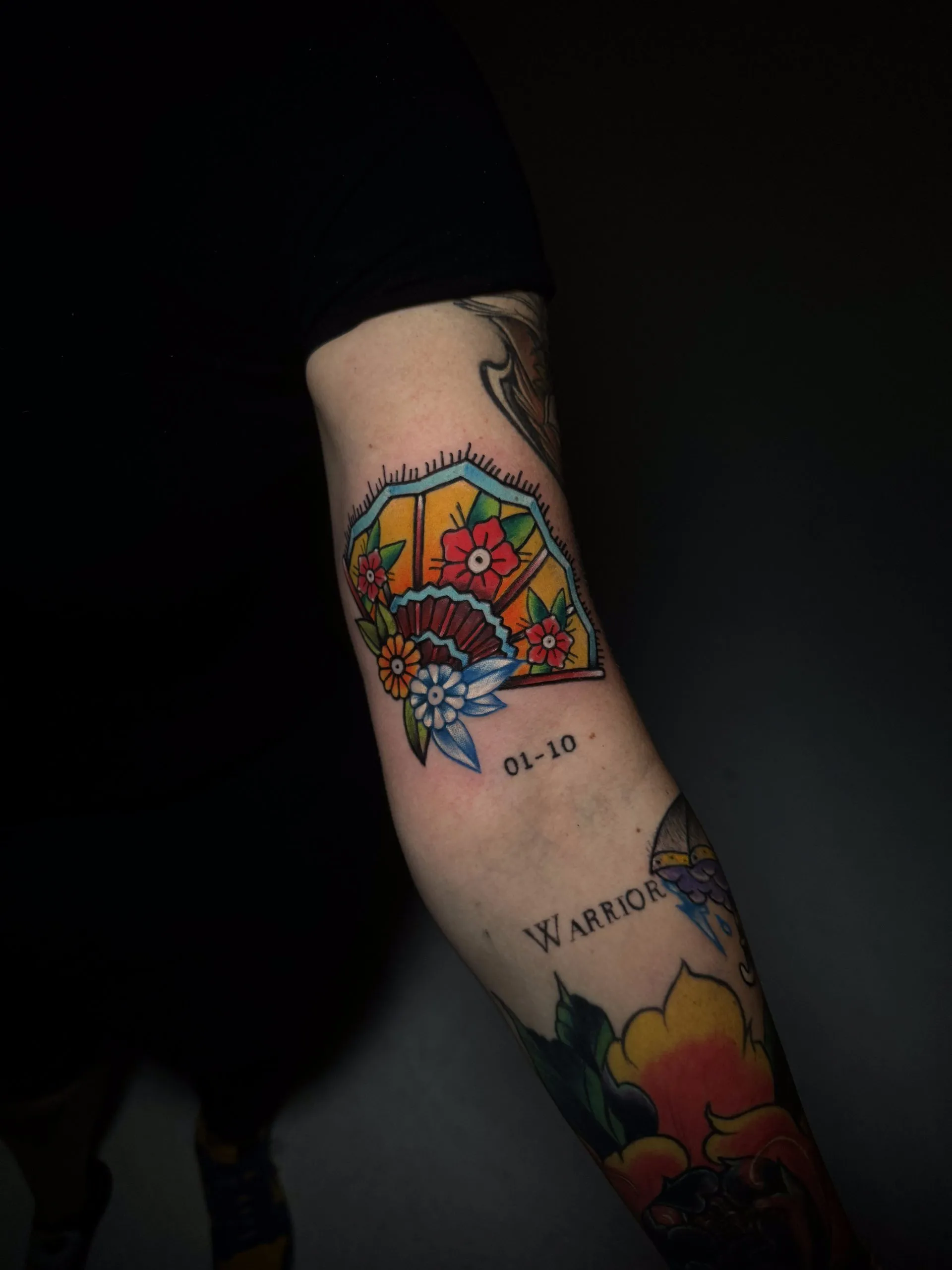 A colorful traditional tattoo on an inner arm. It depicts an open Japanese-style folding fan with a yellow and orange gradient, decorated with several red and blue flowers. The numbers "01-10" and the word "WARRIOR" are tattooed nearby in a simple font.