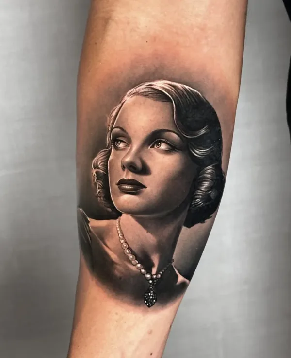 A high-contrast, black-and-grey realism tattoo of a woman’s face in a classic Hollywood style. She is looking upward with a soft expression, featuring wavy hair, a pearl necklace, and detailed shading on her skin and eyes.