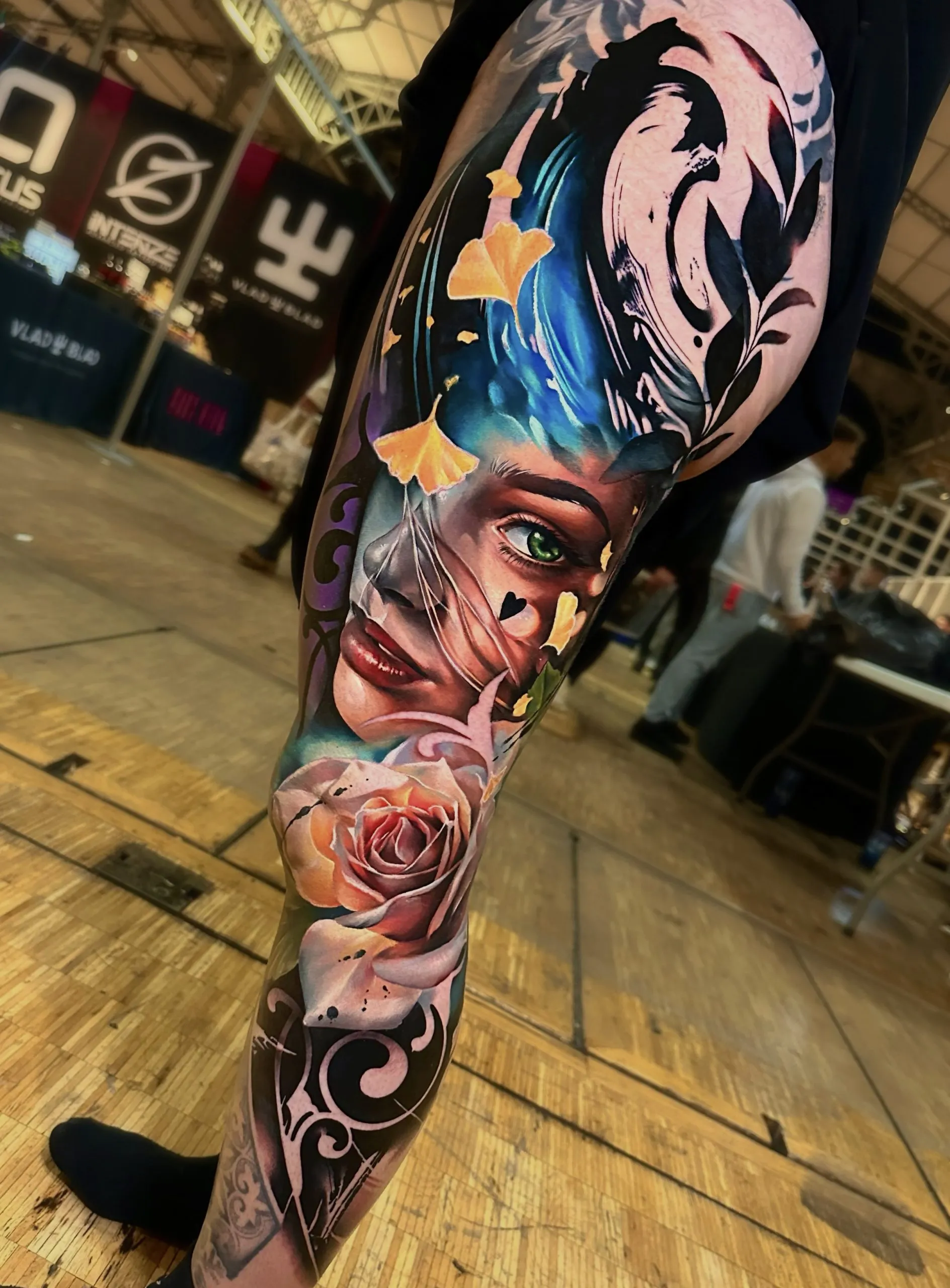 A massive, surrealist color tattoo covering a person's entire leg. The composition blends various elements including a woman's face with a green eye, a large pink rose, falling yellow ginkgo leaves, and bold black abstract flowing patterns over a blue and purple cosmic background.