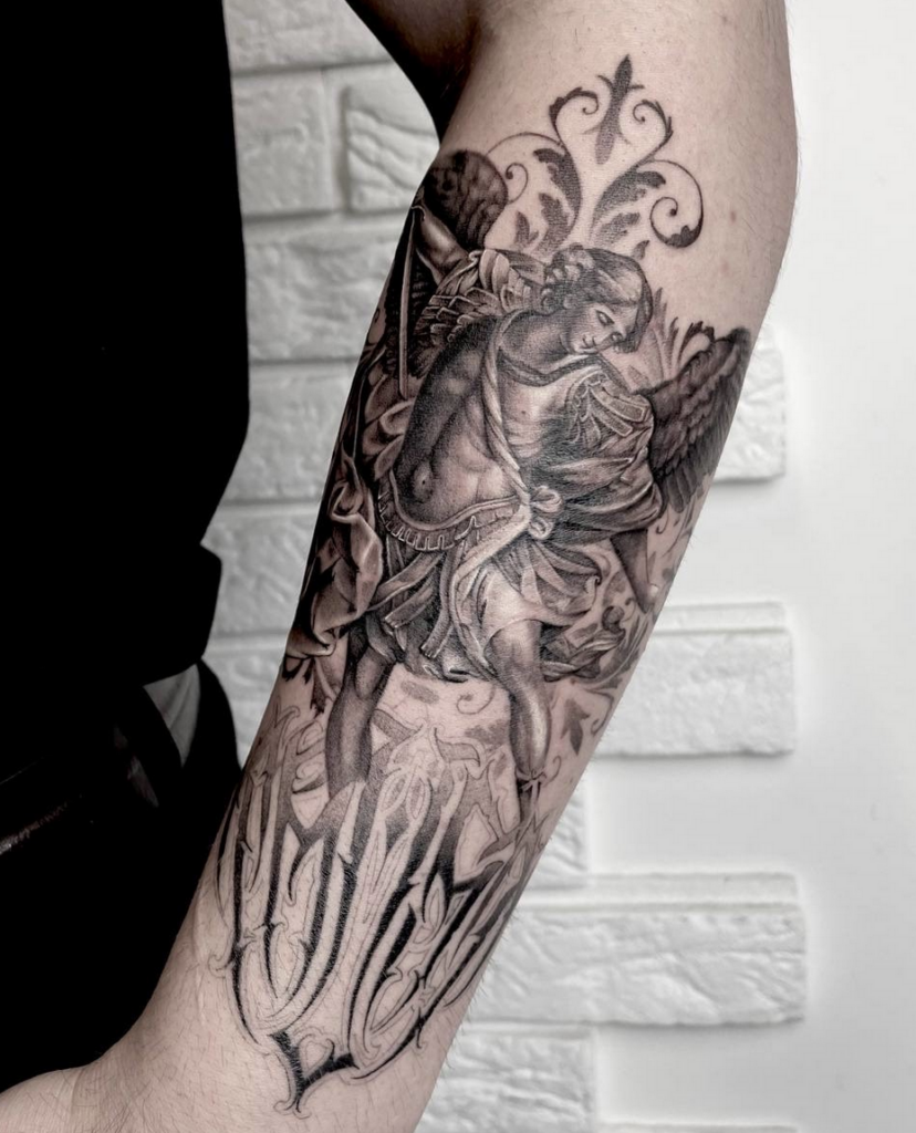 A black-and-grey illustrative tattoo on a person's forearm, depicting a winged angel figure with a sword, executed in a classical style. The angel is set against an ornate filigree background at the top, and bold, ornamental gothic-style script runs along the bottom near the wrist. The background features a textured, light-colored brick wall.