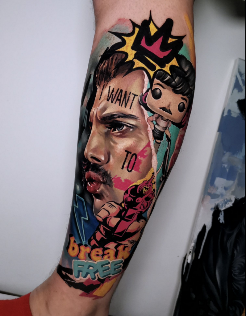 A vibrant, full-color illustrative tattoo on a leg, designed as a pop-art collage. It prominently features a realistic portrait of Freddie Mercury on one side, with a "Funko Pop" style version of the singer in a dress on the other. Red, pink, and turquoise graffiti-style splatters, a pink crown, and a lightning bolt surround the figures. The text "I WANT TO," a pink hair dryer, and the song lyric "break FREE" in mixed block letters are integrated into the dynamic, high-saturation design.
