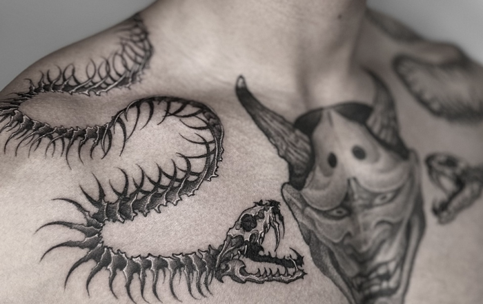 A black-and-white close-up photograph of a person's upper chest and neck, showcasing highly detailed tattoos. On the right side of the chest and wrapping over the shoulder is a long, skeletal snake or centipede-like creature with sharp, curved ribs and a predatory skull for a head. In the center of the chest is a large Hannya mask with horns and a fierce expression. Additional smaller tattoo designs are partially visible on the left side of the chest.
