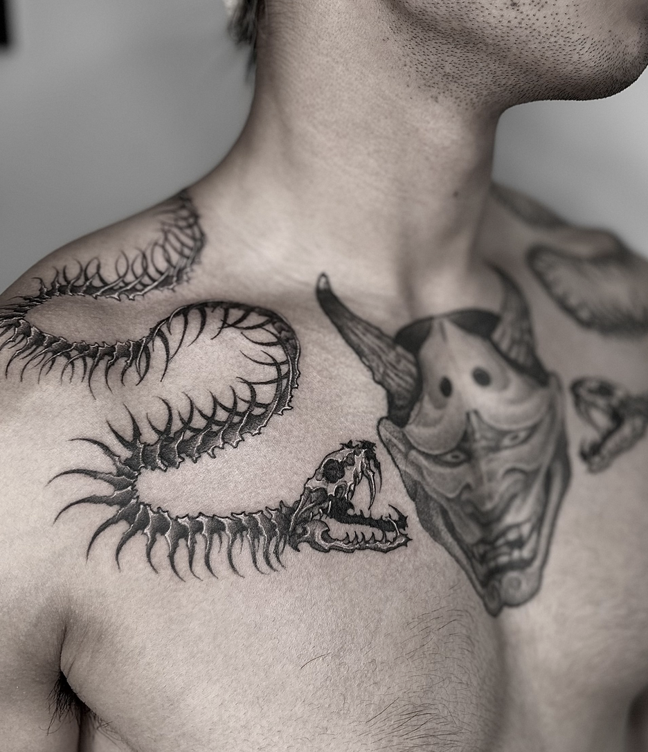 A black-and-white close-up photograph of a person's upper chest and neck, showcasing highly detailed tattoos. On the right side of the chest and wrapping over the shoulder is a long, skeletal snake or centipede-like creature with sharp, curved ribs and a predatory skull for a head. In the center of the chest is a large Hannya mask with horns and a fierce expression. Additional smaller tattoo designs are partially visible on the left side of the chest.