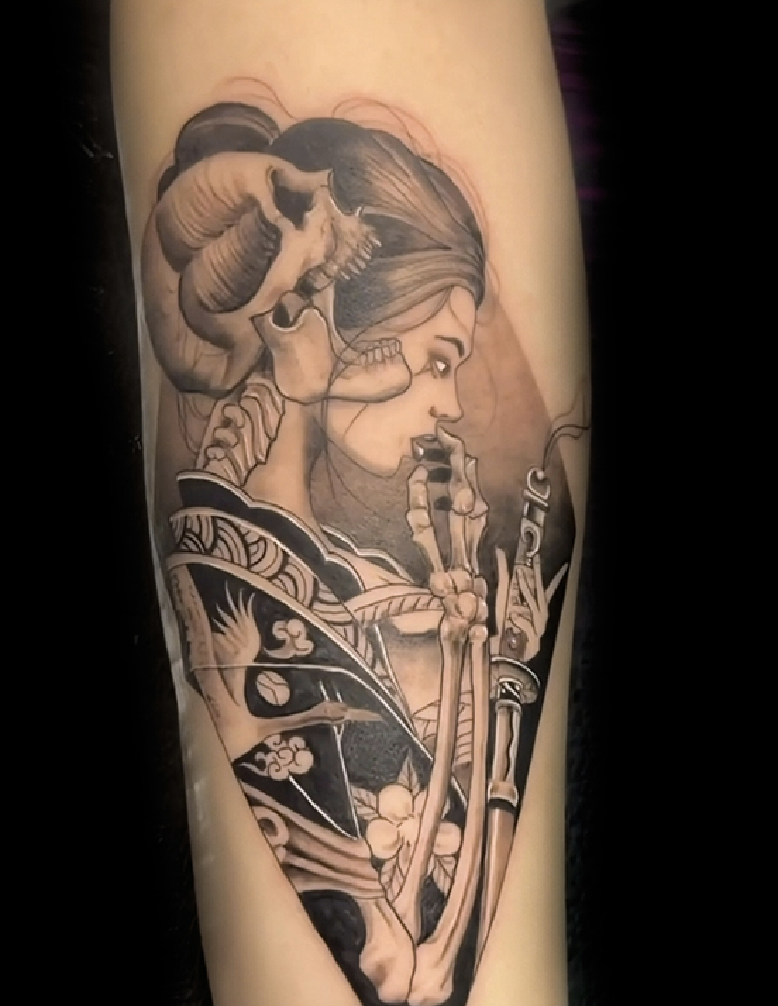 A detailed black-and-grey illustrative tattoo depicting a stylized woman in a traditional patterned kimono holding a katana. Her face is shown in profile, and a large, realistic human skull is integrated into her hair, creating a dark, surreal aesthetic.